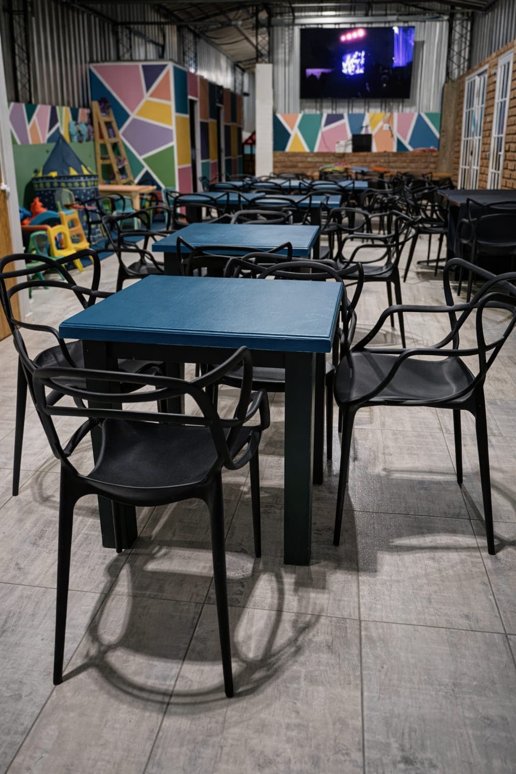 Mesas azules y sillas negras en un salón con murales coloridos y área infantil.