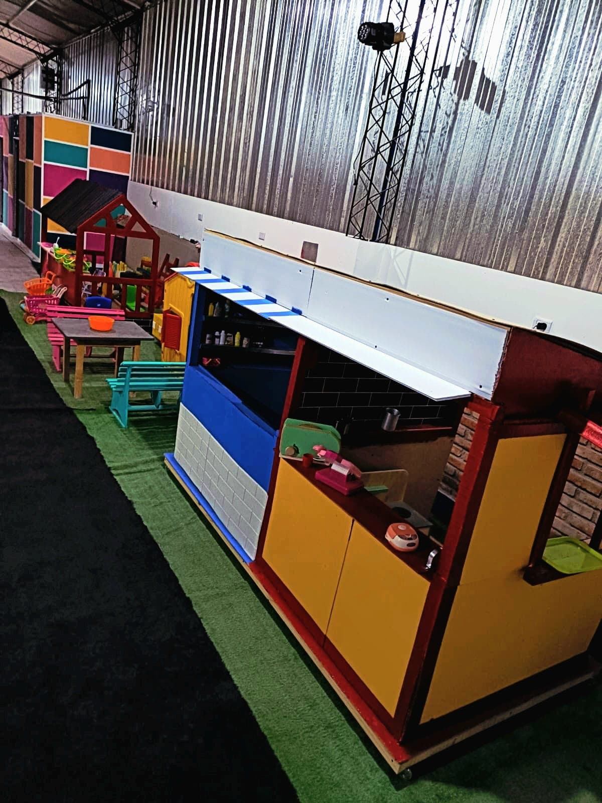 Zona de juegos infantil con puestos de mercado coloridos, casitas de madera y césped artificial.