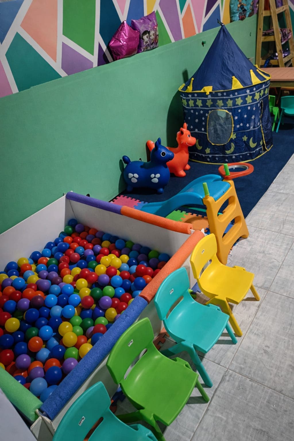 Área de juegos infantil con piscina de pelotas, sillas coloridas, carpa azul y juguetes inflables.