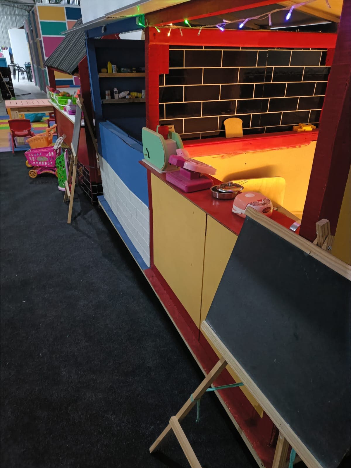 Puesto de mercado infantil con caja registradora, pizarra y carritos de juguete al fondo.