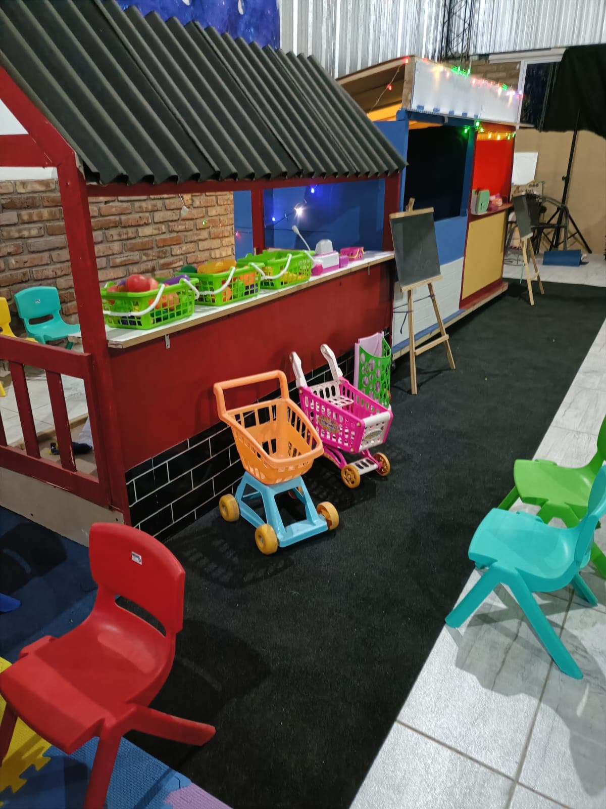 Puesto de mercado infantil rojo con cestas de frutas, carritos de juguete y sillas coloridas.